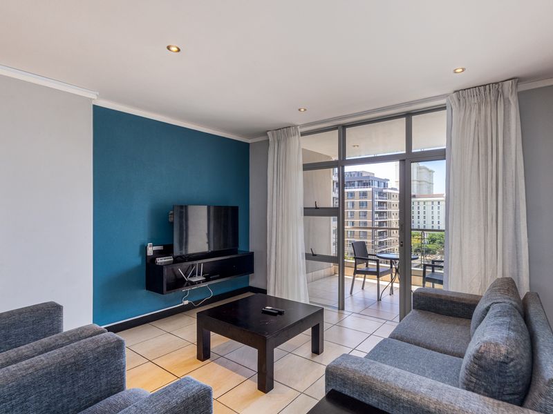 A well-lit living room featuring tiled floors, a bold blue accent wall, and direct access to a private balcony through large sliding glass doors.