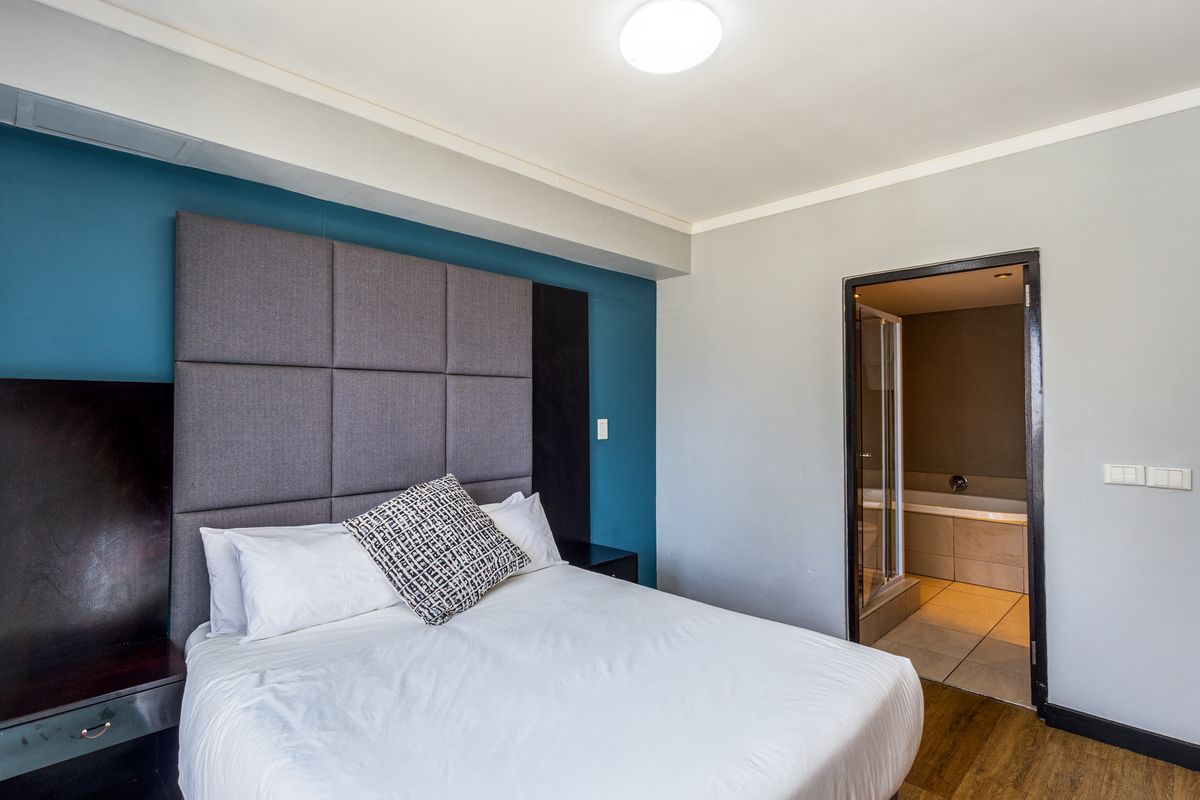 A well-maintained bedroom featuring a modern upholstered headboard, integrated bedside tables, laminate flooring, and direct access to an en-suite bathroom.