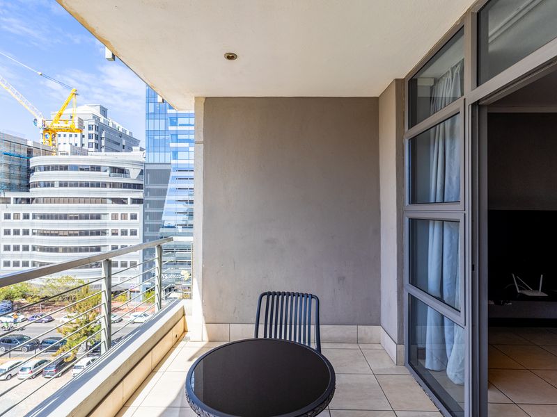 A covered balcony featuring tiled flooring, a metal balustrade, and a sliding glass door, offering an urban view of the surrounding city buildings.