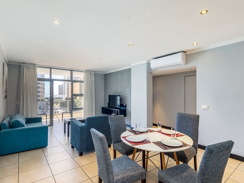 A bright open-plan living and dining area featuring tiled floors, recessed lighting, a wall-mounted air conditioning unit, and access to a private balcony.