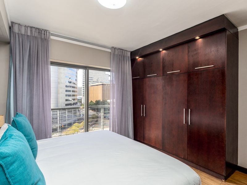 A well-maintained bedroom featuring dark wood built-in cupboards with integrated lighting, laminate flooring, and a sliding door leading to a balcony with city views.