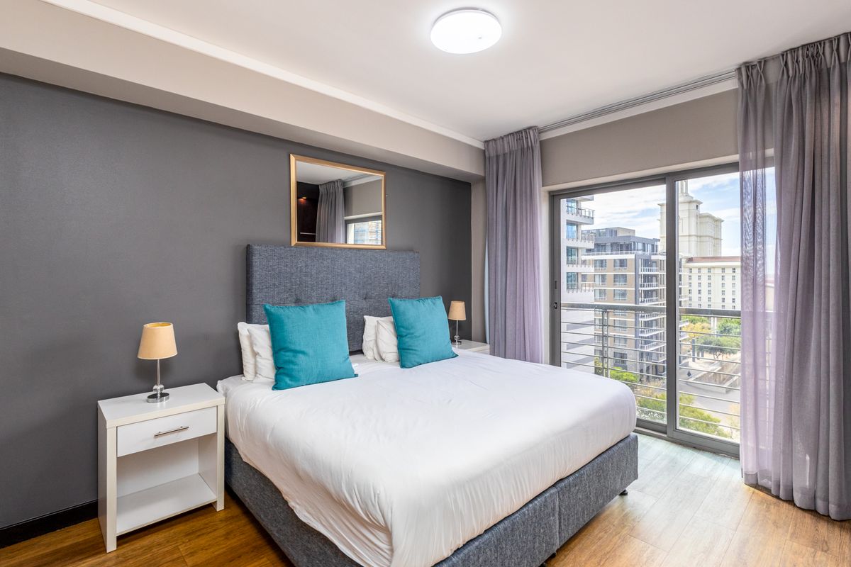 A well-maintained bedroom featuring laminate flooring, a feature wall, and a sliding glass door providing access to a private balcony with city views.