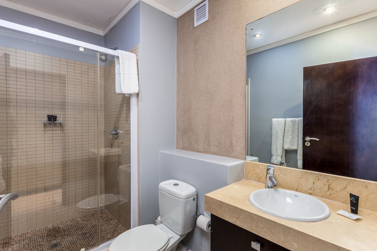 A functional bathroom featuring a glass-enclosed shower with mosaic tiling, a stone-topped vanity with an undermount sink, and a large wall-mounted mirror.