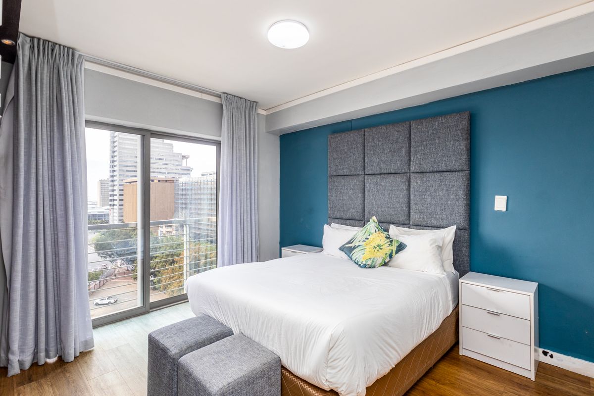 A well-maintained bedroom featuring laminate flooring, a bold blue accent wall, and a sliding glass door providing access to a private balcony.