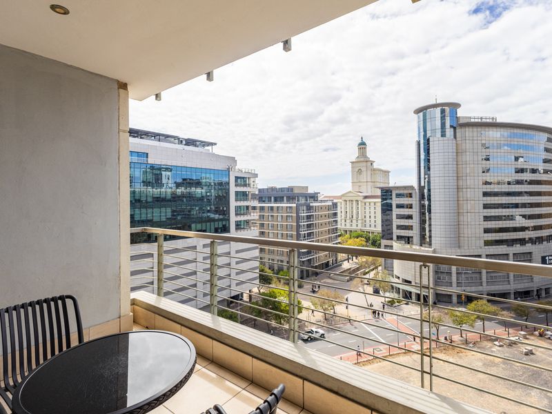 A covered balcony featuring tiled flooring and a stainless steel balustrade, offering an elevated urban view of the surrounding city buildings.