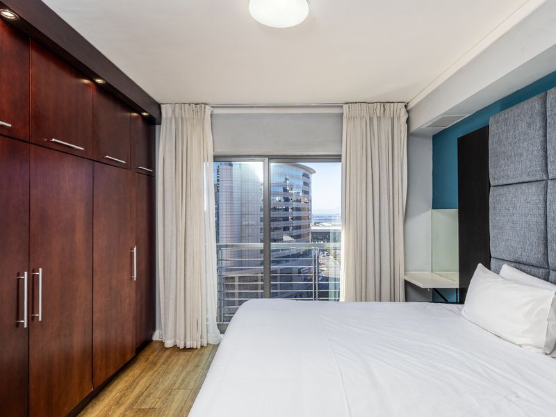 A well-maintained bedroom featuring extensive built-in wooden cupboards, laminate flooring, and a sliding door leading to a balcony with city views.