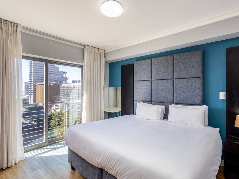A well-maintained bedroom featuring a built-in upholstered headboard, laminate flooring, and a sliding glass door leading to a balcony with city views.