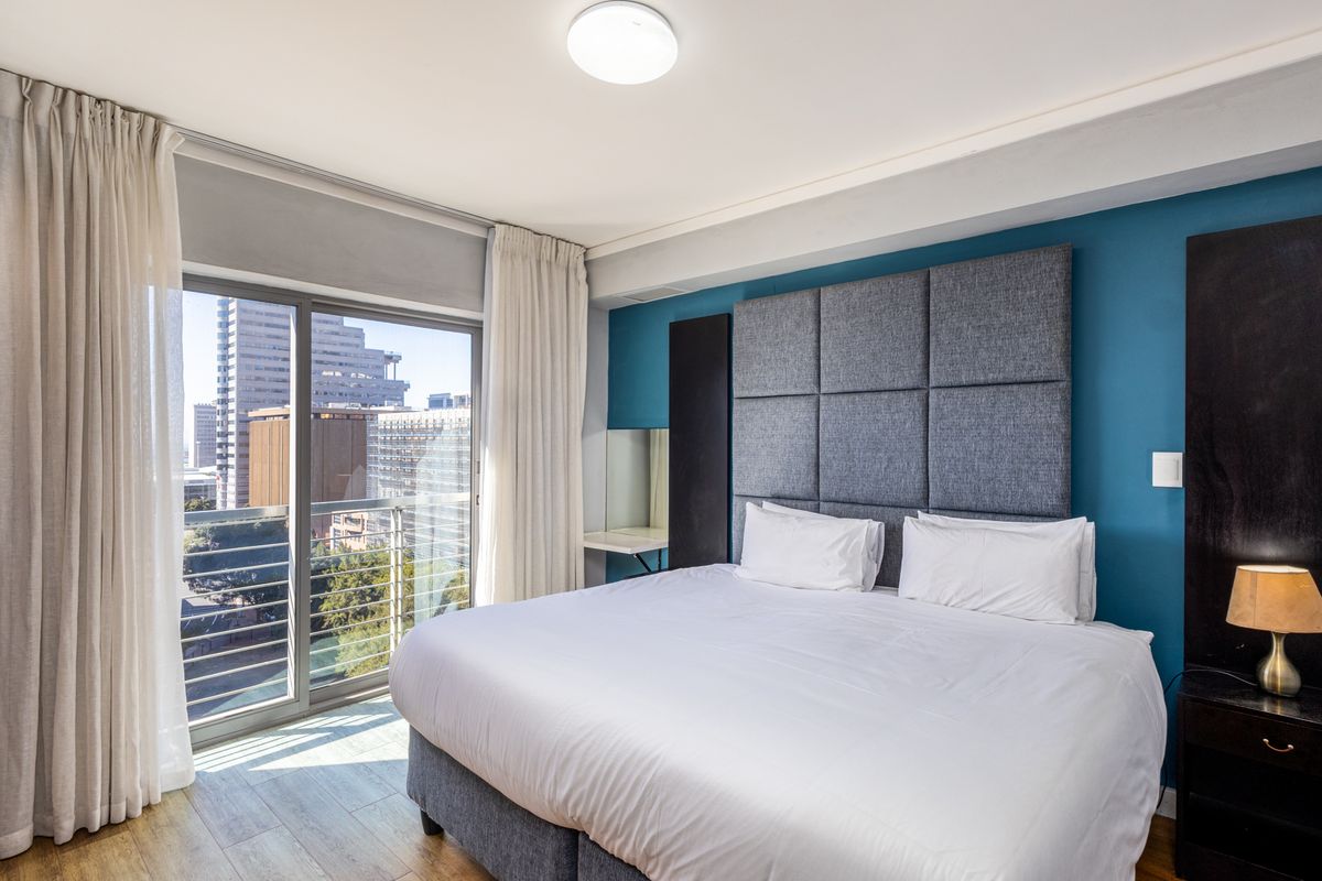 A well-maintained bedroom featuring a built-in upholstered headboard, laminate flooring, and a sliding glass door leading to a balcony with city views.