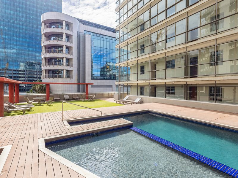 A well-maintained outdoor pool area featuring timber decking, a modern swimming pool, and a red metal pergola set against a contemporary apartment building.