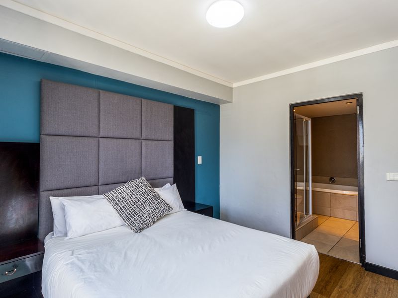 A well-maintained bedroom featuring a modern upholstered headboard, integrated bedside tables, laminate flooring, and direct access to an en-suite bathroom.