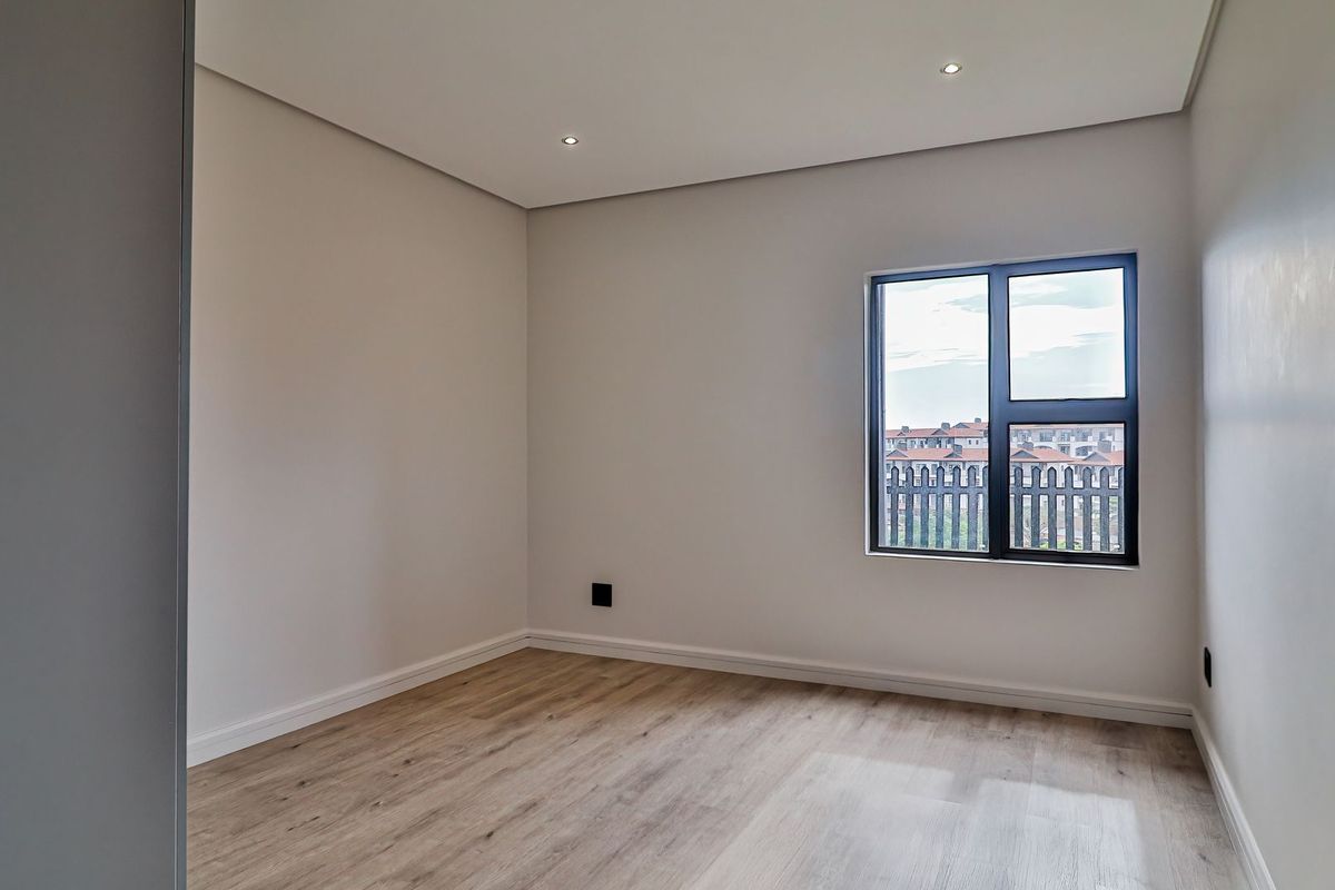 A bright, empty bedroom featuring modern wood-look flooring, neutral walls, recessed ceiling lighting, and a large window with dark framing.