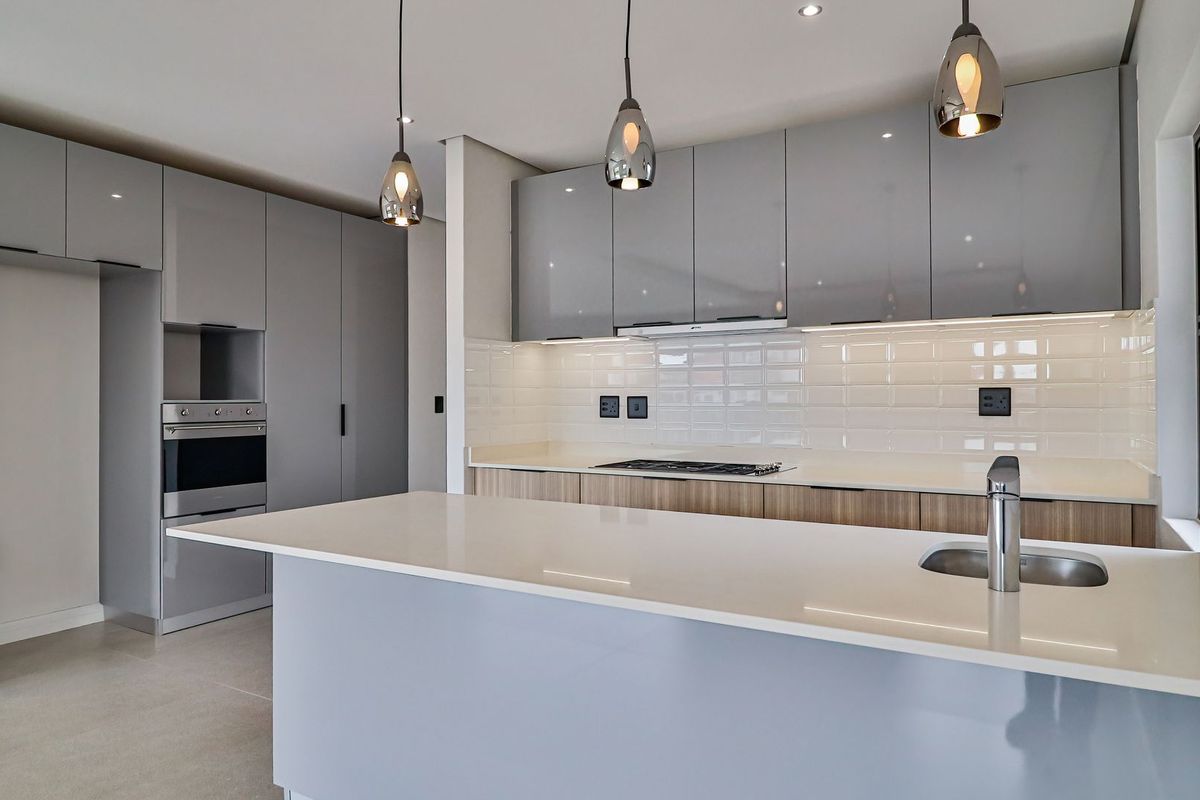 Modern kitchen featuring high-gloss grey cabinetry, stone countertops, a central island with a sink, built-in oven, and subway tile backsplash.