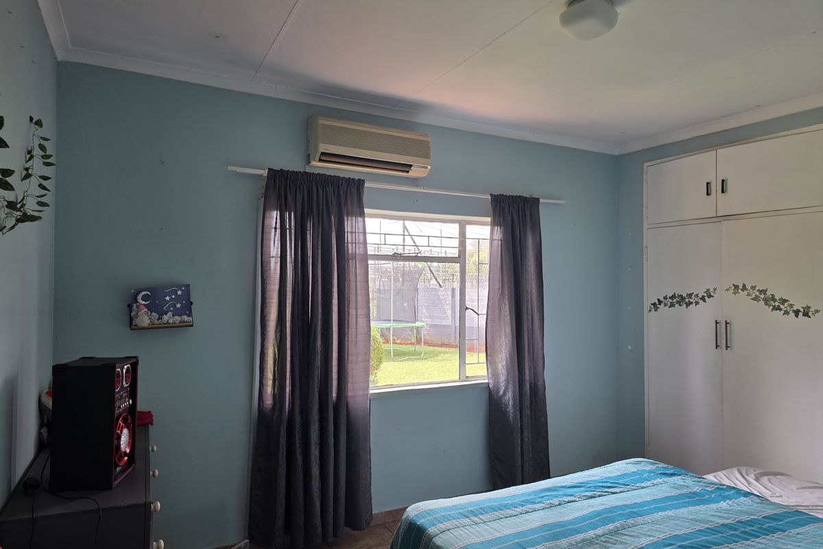 A bedroom featuring built-in cupboards, tiled flooring, and an air conditioning unit, with natural light provided by a window with security bars.
