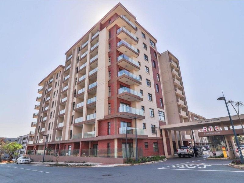 A modern multi-story apartment building featuring brick and plaster facade, glass-railed balconies, and a secure gated entrance with a covered driveway.