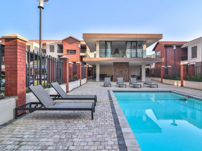 Communal swimming pool area featuring paved decking, brick boundary walls with metal fencing, and a modern clubhouse structure in the background.