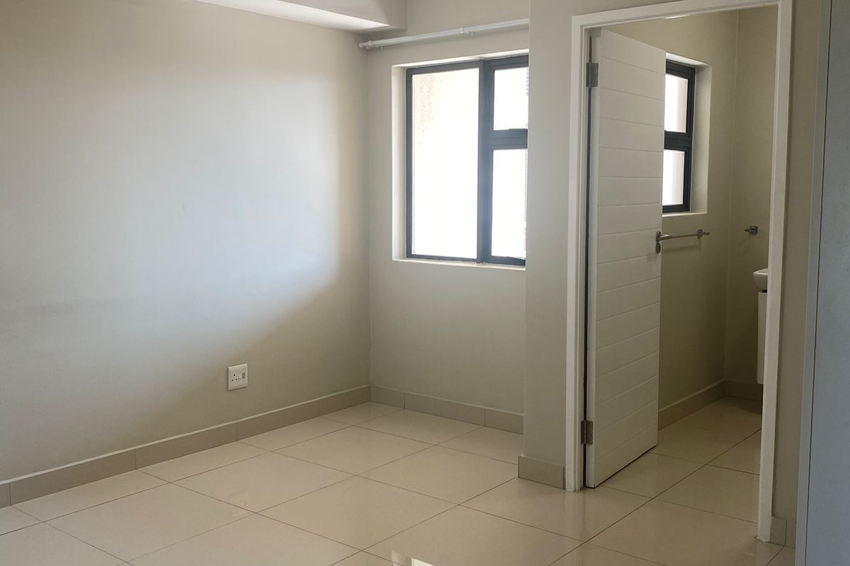 A bright, empty bedroom featuring neutral tiled flooring, modern ceiling lighting, and an adjoining en-suite bathroom with a clean, minimalist finish.
