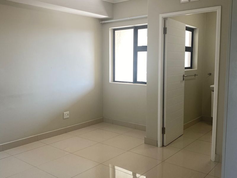 A bright, empty bedroom featuring neutral tiled flooring, modern ceiling lighting, and an adjoining en-suite bathroom with a clean, minimalist finish.