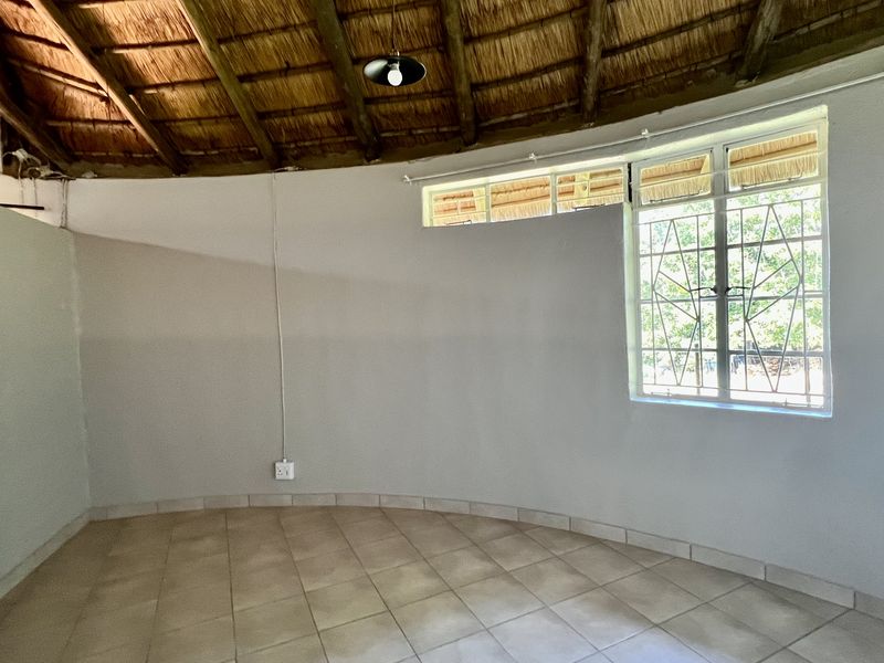 An empty living area featuring a traditional thatched roof with exposed timber rafters, tiled flooring, and a large window with security burglar bars.