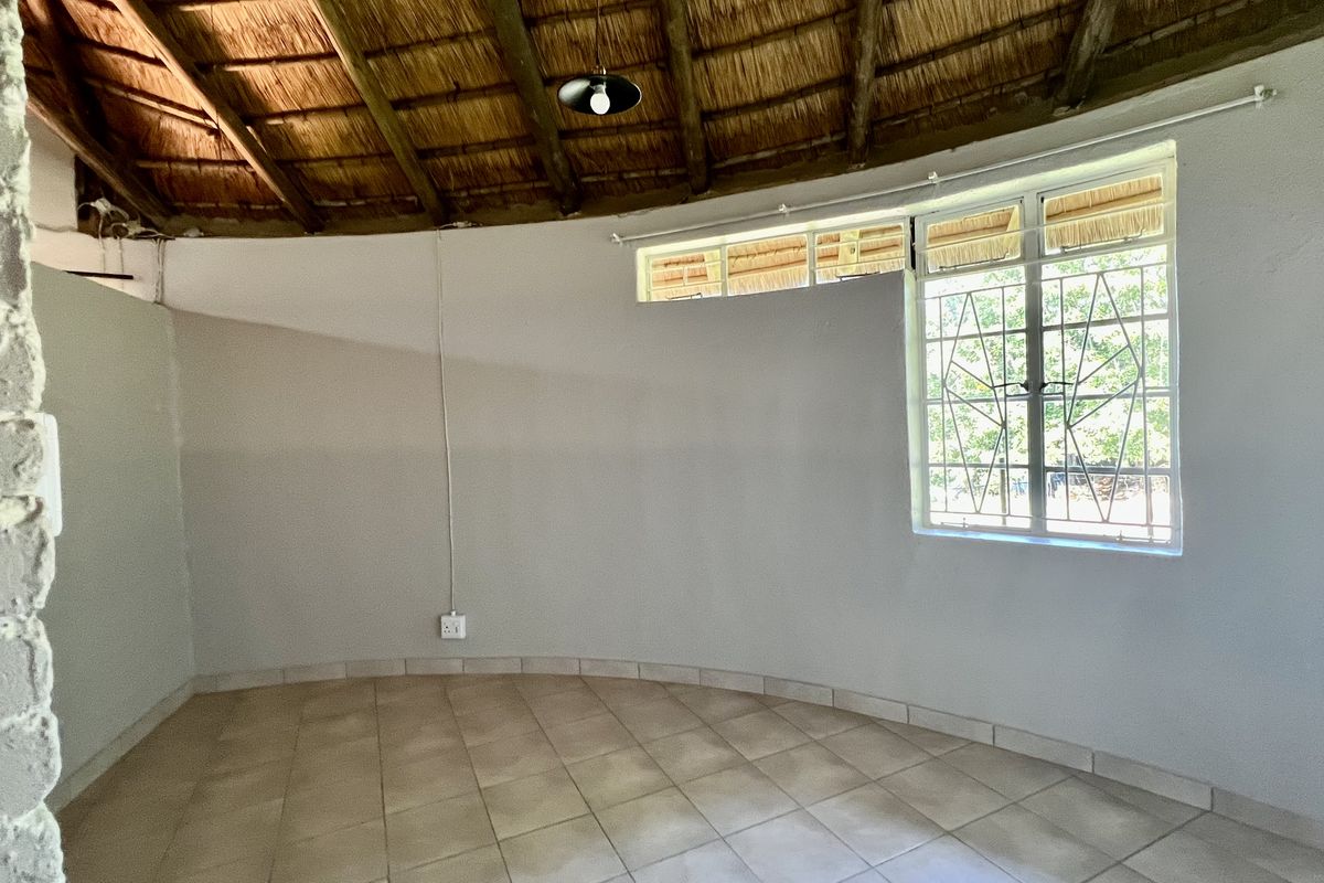 An empty living area featuring a traditional thatched roof with exposed timber rafters, tiled flooring, and a large window with security burglar bars.