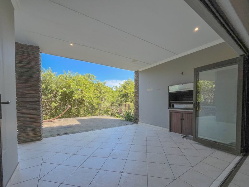 A spacious covered patio featuring tiled flooring, stone-clad pillars, and a built-in braai, leading out to an elevated wooden deck with garden views.