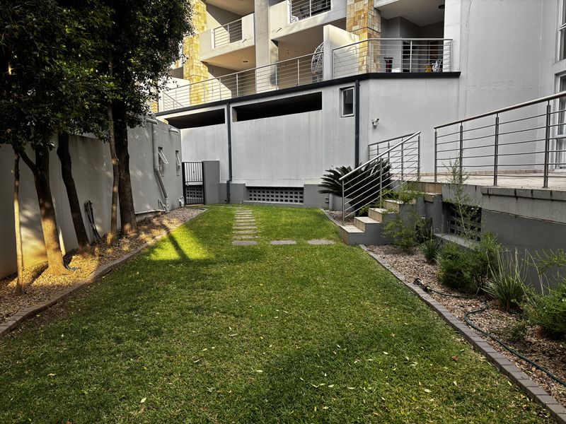 A private rear garden area featuring a manicured lawn, stone stepping path, and concrete stairs leading up to the apartment balcony.