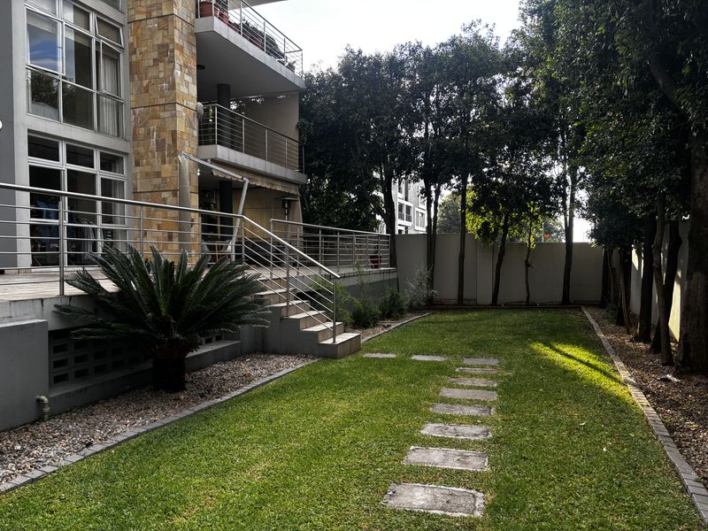 A well-maintained rear garden featuring a manicured lawn, stone stepping path, and direct access to the apartment's elevated patio via a stainless steel staircase.