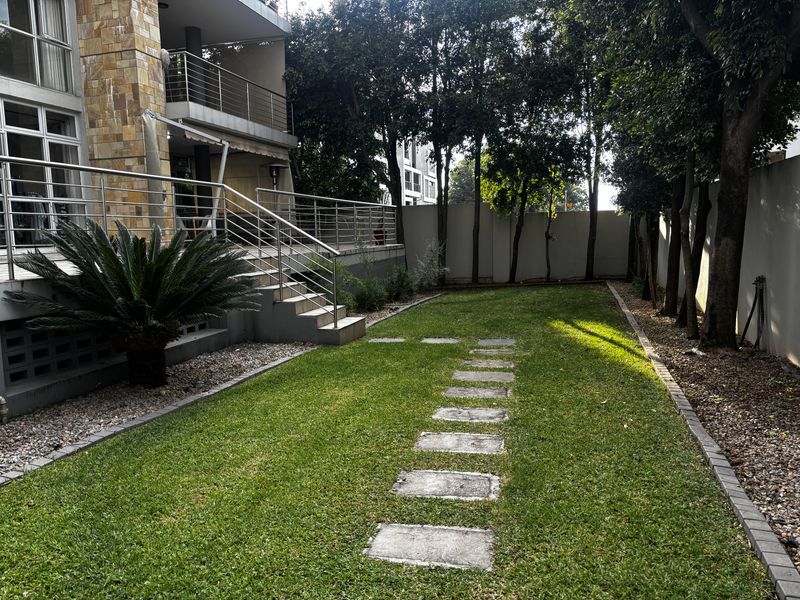 A well-maintained rear garden featuring a manicured lawn, stone paver pathway, and a raised patio area with stainless steel railings.