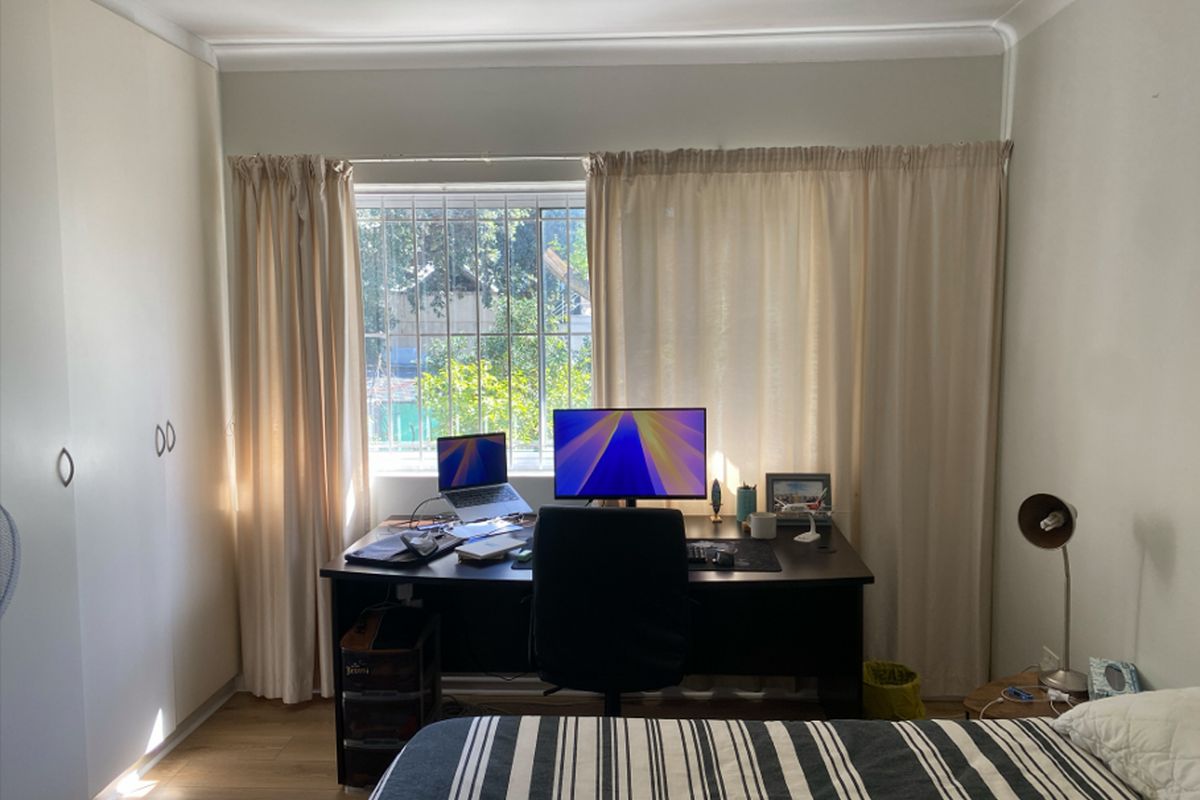 A functional bedroom featuring built-in cupboards, laminate flooring, and a window with security bars, currently utilized as a home office space.