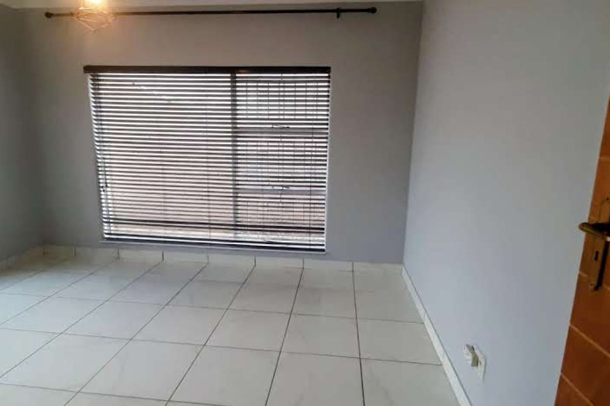 A bright, empty bedroom featuring neutral tiled flooring, large windows with venetian blinds, and a solid wooden door.