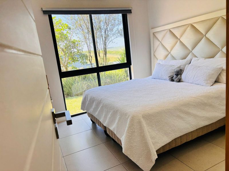 A bright bedroom featuring large aluminium-framed windows with garden views, neutral tiled flooring, and a modern aesthetic.