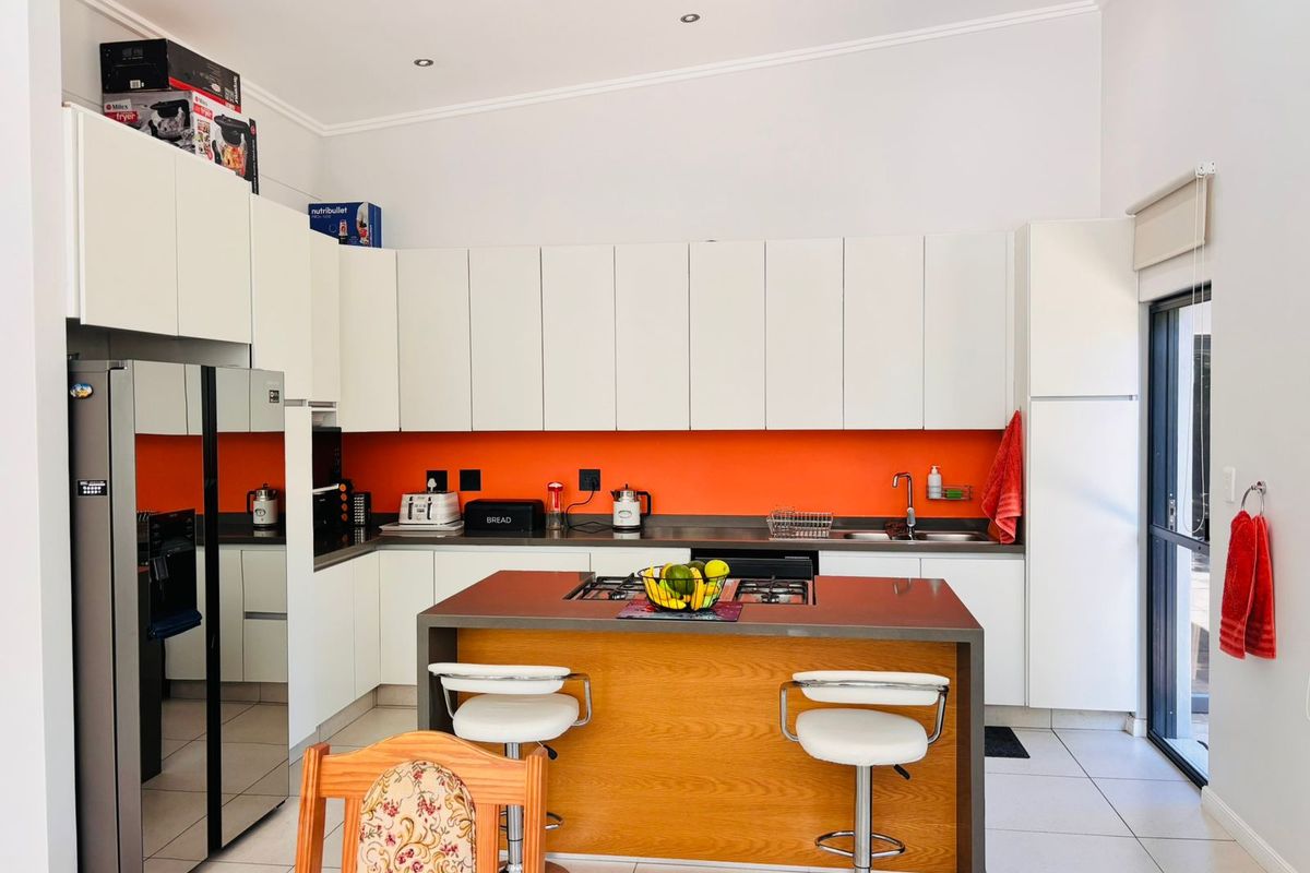 Modern kitchen featuring white cabinetry, a central island with a gas hob, vibrant orange splashback, and tiled flooring with direct outdoor access.