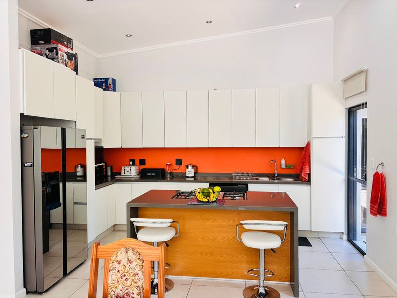 Modern kitchen featuring white cabinetry, a central island with a gas hob, vibrant orange splashback, and tiled flooring with direct outdoor access.