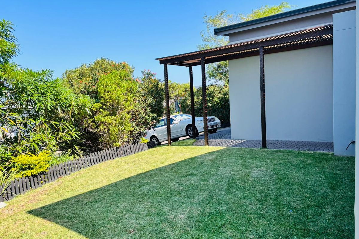 A well-maintained rear garden area featuring a modern pergola structure, paved driveway section, and established landscaping with a picket fence.