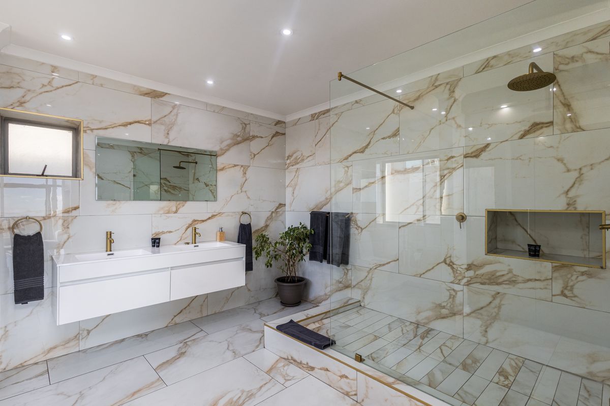 Modern bathroom featuring a double vanity, walk-in shower with glass screen, rain shower head, and elegant marble-look tiling with brass accents.