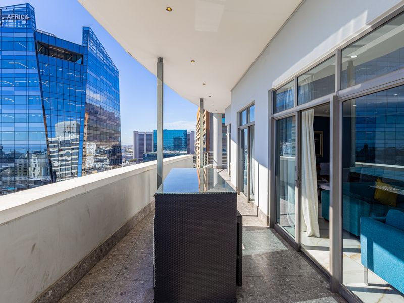 A spacious covered balcony featuring stone flooring, recessed lighting, and sliding glass doors, offering an elevated urban view of the surrounding city skyline.
