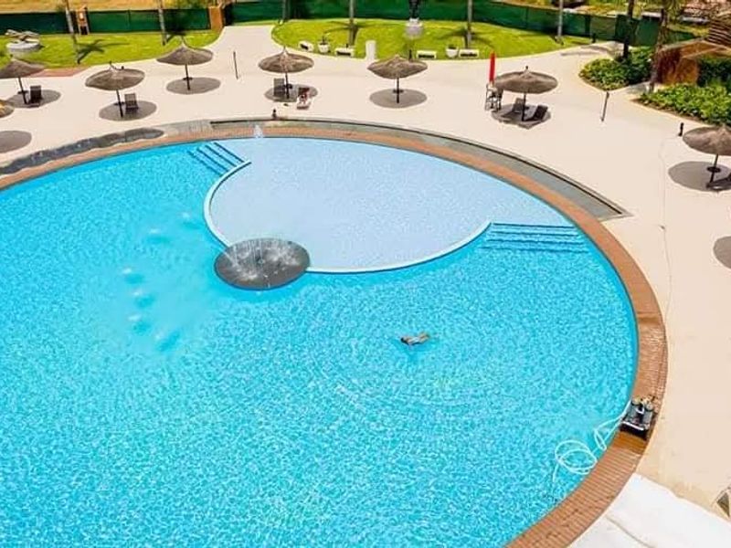 Aerial view of a large circular swimming pool with a central fountain feature, surrounded by a paved deck and landscaped garden area.