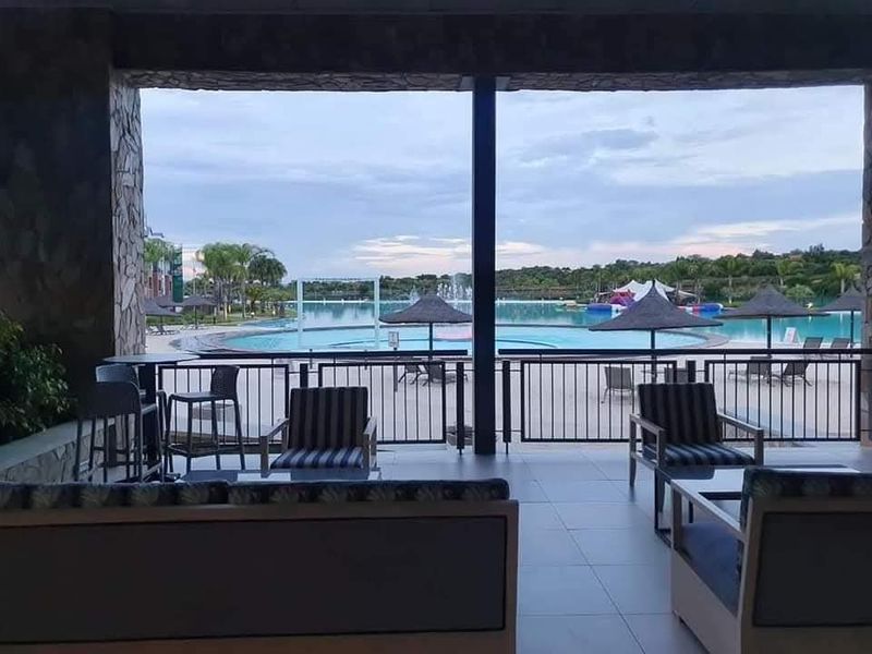 A covered patio area overlooking a large communal swimming pool and landscaped grounds, featuring tiled flooring and stone-clad structural pillars.