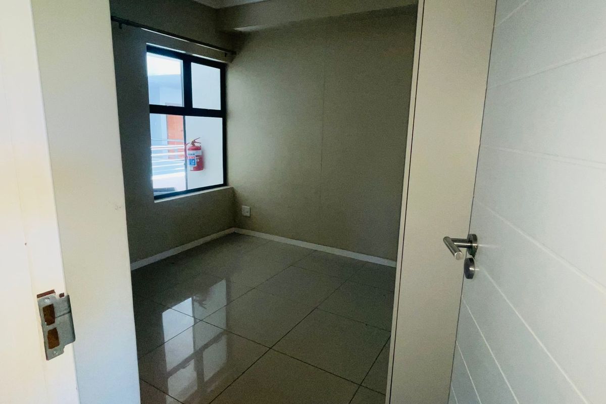 A compact, empty bedroom featuring neutral wall tones, large format floor tiles, and a window providing natural light.