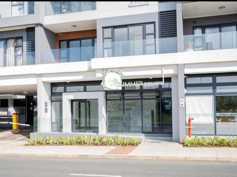 Modern apartment building exterior featuring glass balcony railings, large aluminum-framed windows, and ground-level commercial frontage with paved access.