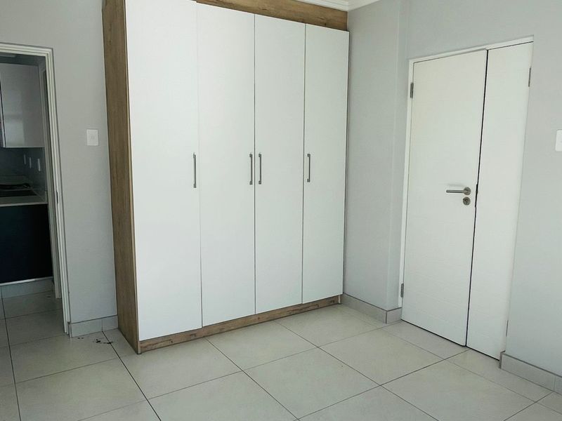 A bright bedroom featuring modern built-in cupboards with a wood-look frame, neutral floor tiles, and direct access to an adjacent room.