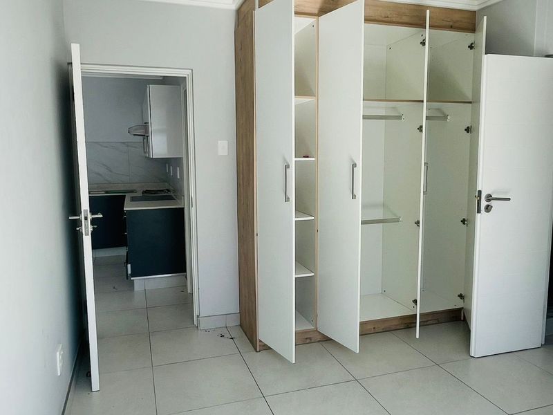 A bright bedroom featuring built-in wooden cabinetry with white doors and tiled flooring, with an adjacent view into a modern kitchen area.