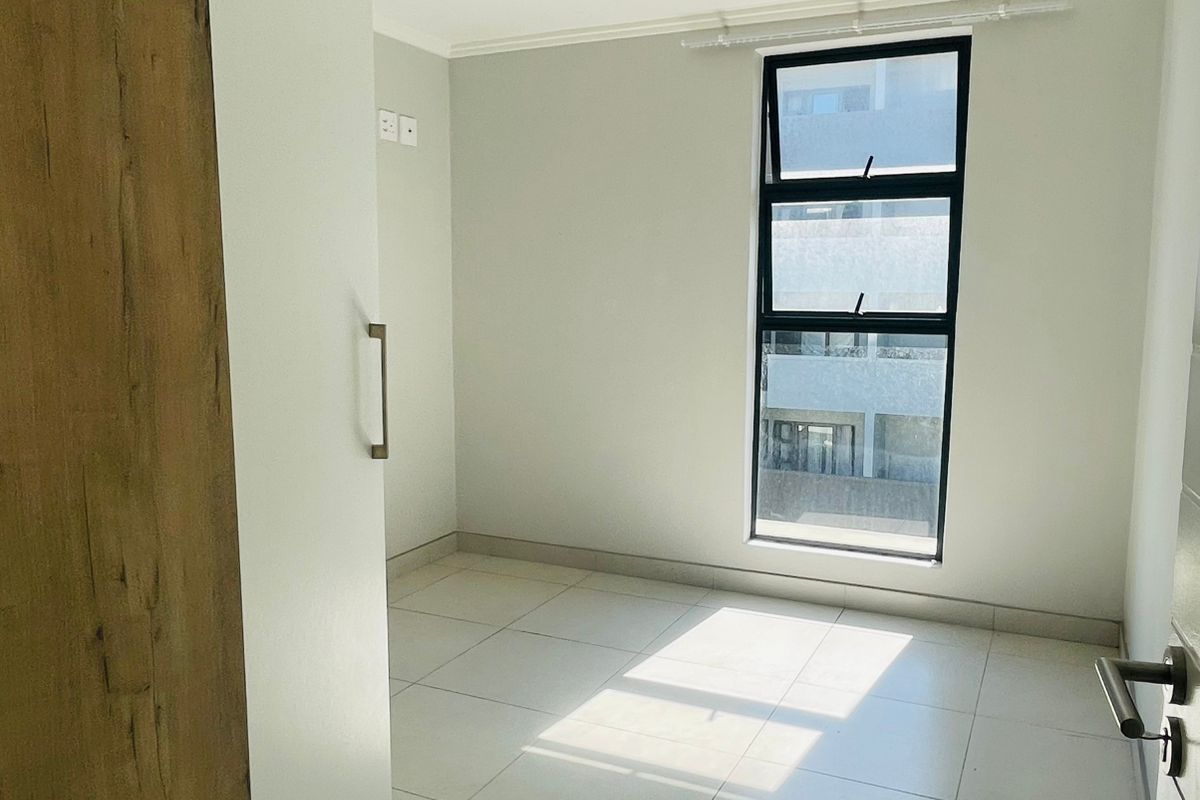 A bright, empty bedroom featuring neutral tiled flooring, a modern black-framed window, and built-in cabinetry with a wood-grain finish.