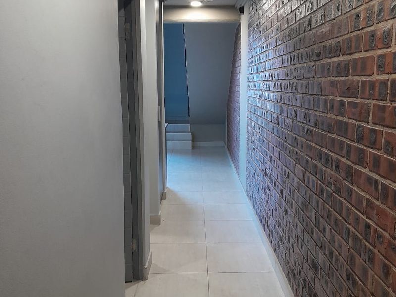 A well-lit hallway featuring a distinctive exposed brick feature wall, tiled flooring, and recessed ceiling lighting leading towards a staircase.