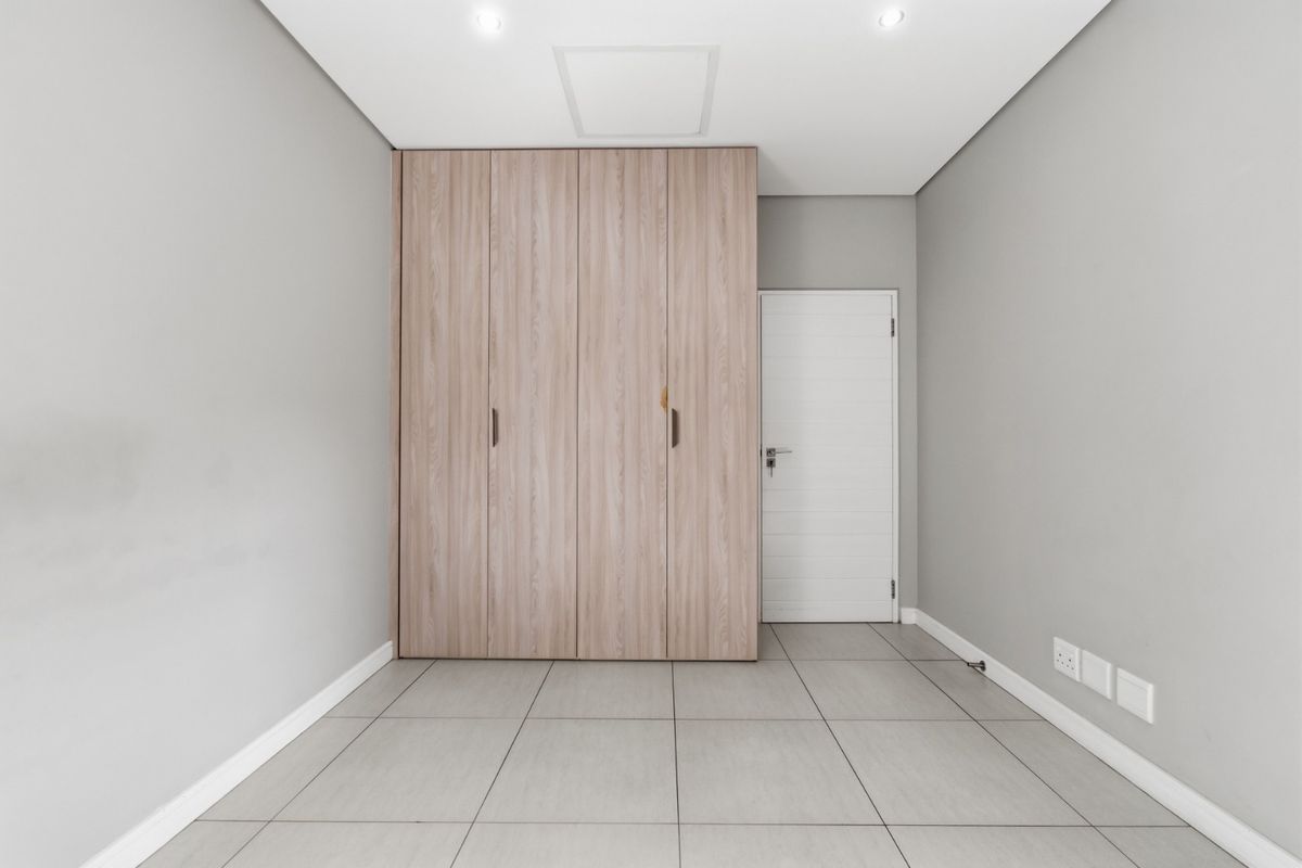 A clean, modern bedroom featuring neutral grey walls, large format floor tiles, and built-in light wood-grain cabinetry.