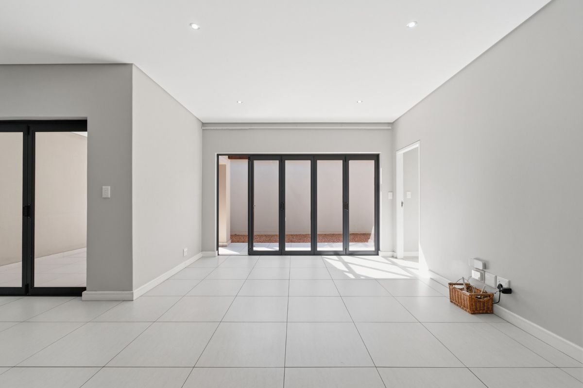 A bright, modern, and spacious living area featuring neutral tiled flooring, recessed ceiling lighting, and large dark-framed sliding glass doors leading to the exterior.