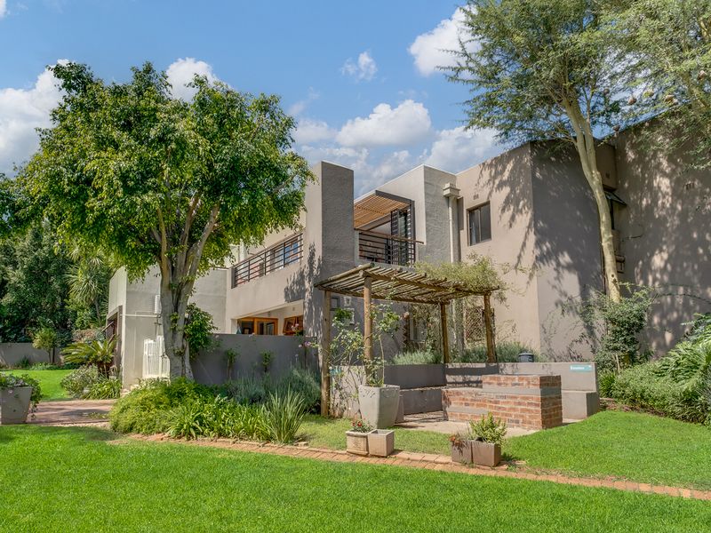 Rear view of the property featuring a landscaped garden, a brick built-in braai area with a wooden pergola, and an upper-level balcony.