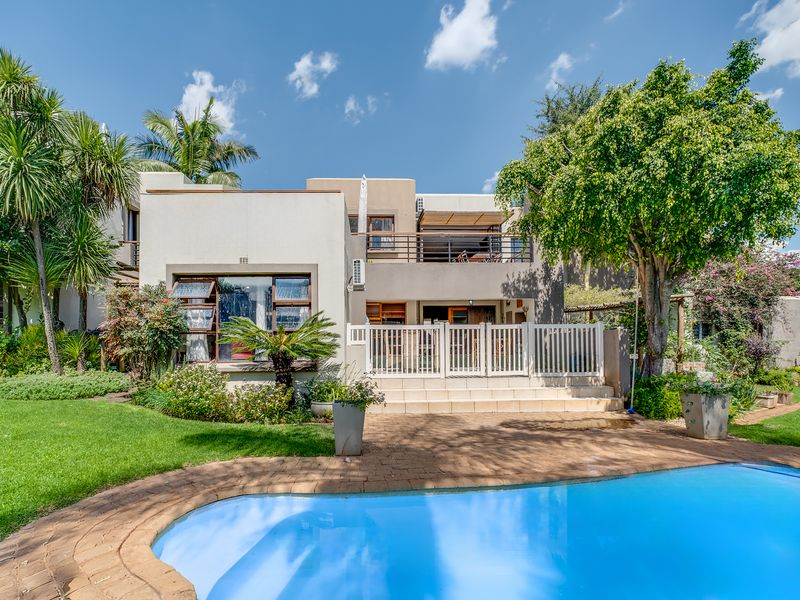Rear view of the property featuring a swimming pool, paved patio area, and an upper-level balcony overlooking the landscaped garden.