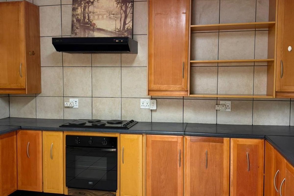 A functional kitchen featuring wooden cabinetry, dark countertops, a built-in oven, and a black extractor fan with tiled splashbacks and flooring.
