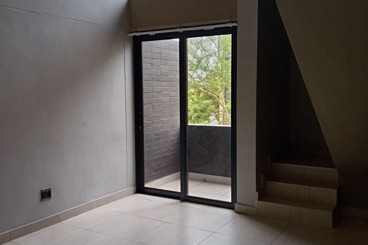 A bright, double-volume living area featuring tiled flooring, a staircase, and large sliding glass doors leading to a private balcony.