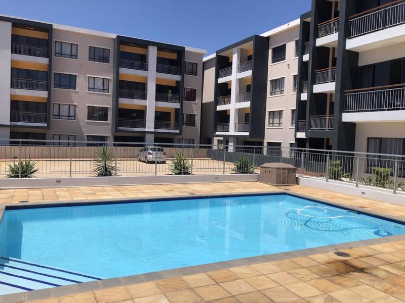A well-maintained communal swimming pool area featuring a tiled deck and metal fencing, set against the backdrop of a modern multi-story apartment complex.